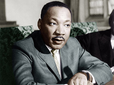 A man in a suit sits at a table, looking thoughtful. His posture is attentive, and he has a mustache. The background shows blurred interior details.